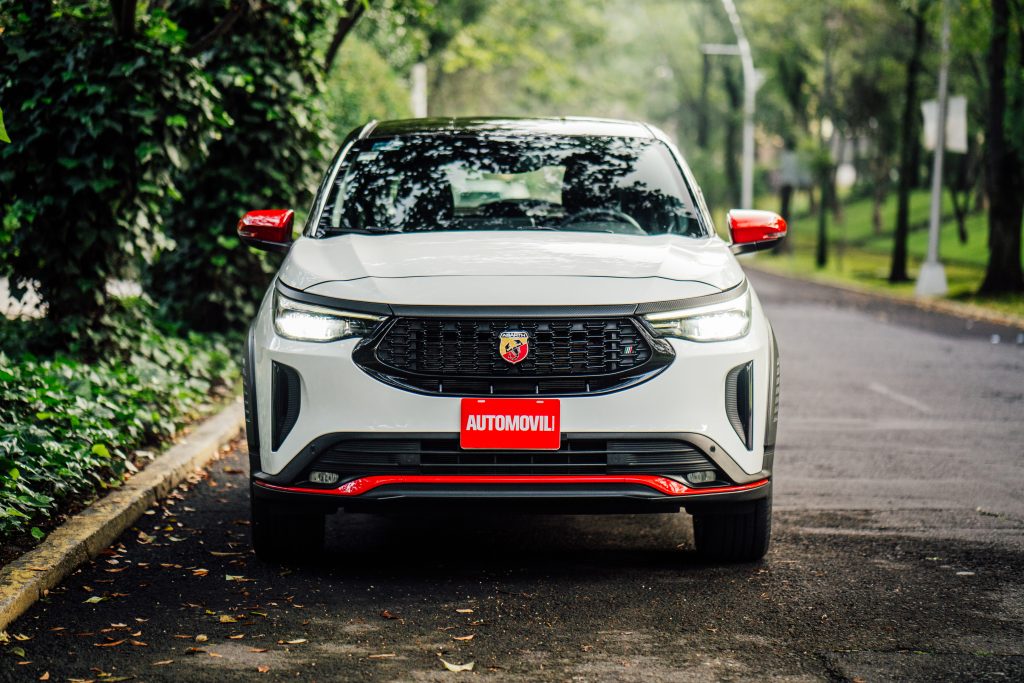 Una SUV Fiat Pulse Abarth blanca con detalles rojos conduciendo por una calle arbolada. La parte delantera del automóvil muestra el logotipo de Abarth y la palabra "AUTOMOVIL" en la matrícula.