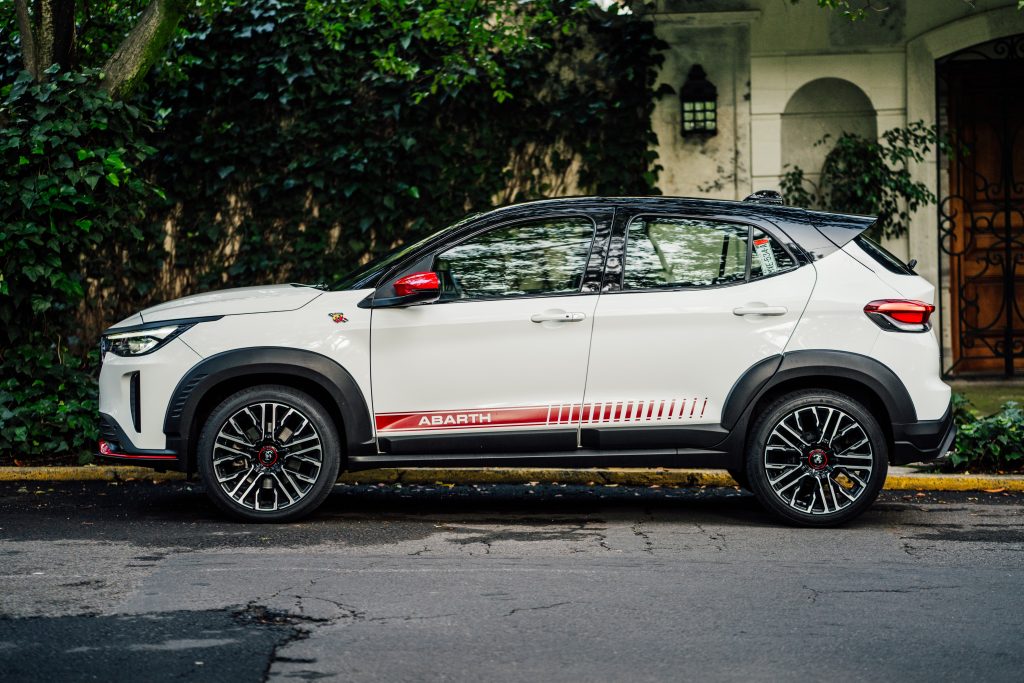 Vista lateral de un SUV compacto Abarth Pulse de color blanco perlado con techo negro, rines deportivos negros y franjas rojas características de la marca. El vehículo está estacionado en una calle arbolada con una casa de estilo clásico en el fondo.