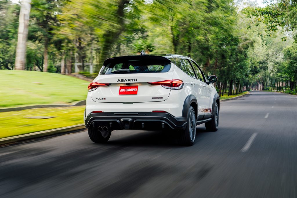 Un Abarth Pulse blanco, modelo SUV compacto, es capturado desde atrás mientras acelera por una carretera rodeada de árboles. Destaca el diseño deportivo del vehículo, con un difusor trasero y doble salida de escape.