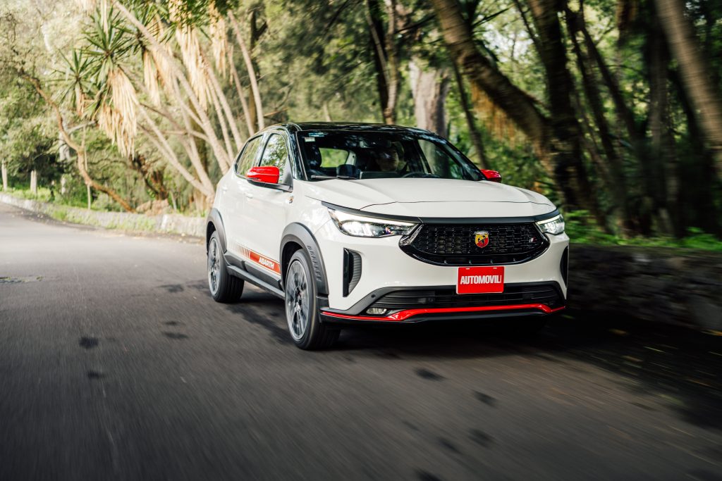 Un Fiat Pulse Abarth blanco en movimiento, desplazándose por una carretera rodeada de árboles. El vehículo muestra detalles en rojo, como los espejos laterales y la parrilla frontal, resaltando su diseño deportivo.